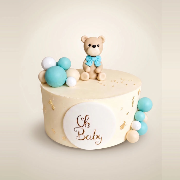 Tort na baby shower dla chłopca - kremowy tort z misiem z masy cukrowej z błękitną kokardą, pastelowymi kulami, złotymi płatkami i plakietką Oh Baby