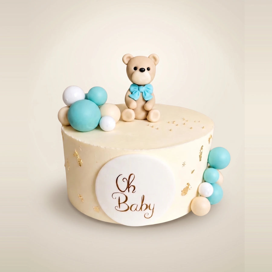 Tort na baby shower dla chłopca - kremowy tort z misiem z masy cukrowej z błękitną kokardą, pastelowymi kulami, złotymi płatkami i plakietką Oh Baby